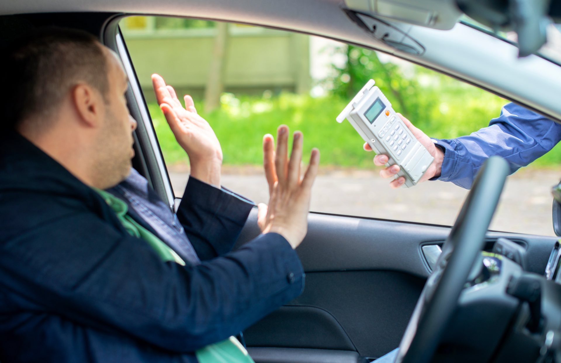 Person sitting in a car with his hands up and another hand outside the window holding a breathalyzer