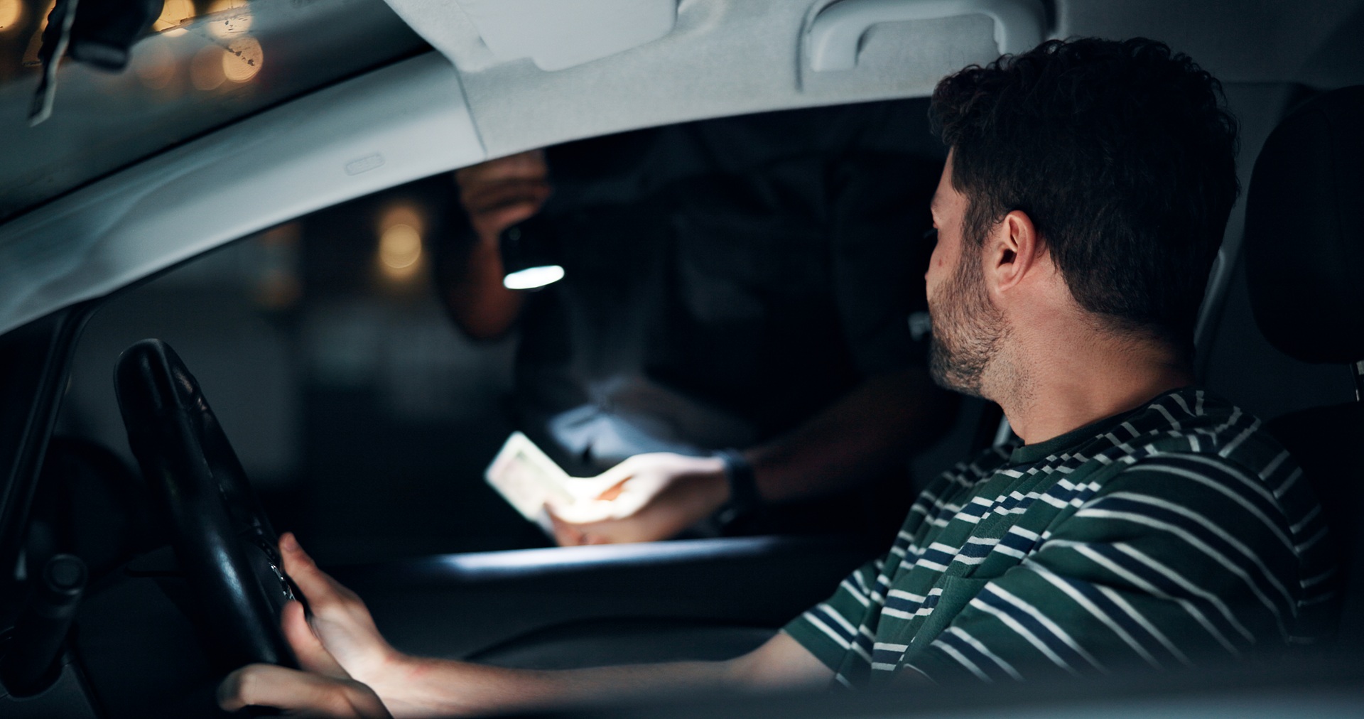 A person pulled over in the dark sitting in the drivers seat while a police officer looks at paperwork under a flashlight.